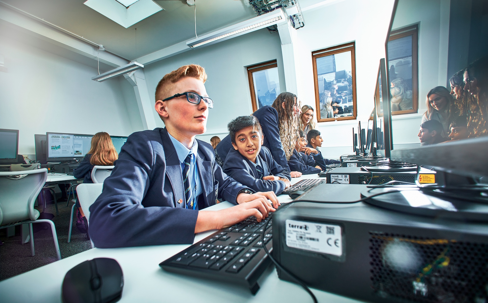 Five students and a teacher sitting around computer screens collaborating with a boy in the middle looking straight down the camera smiling