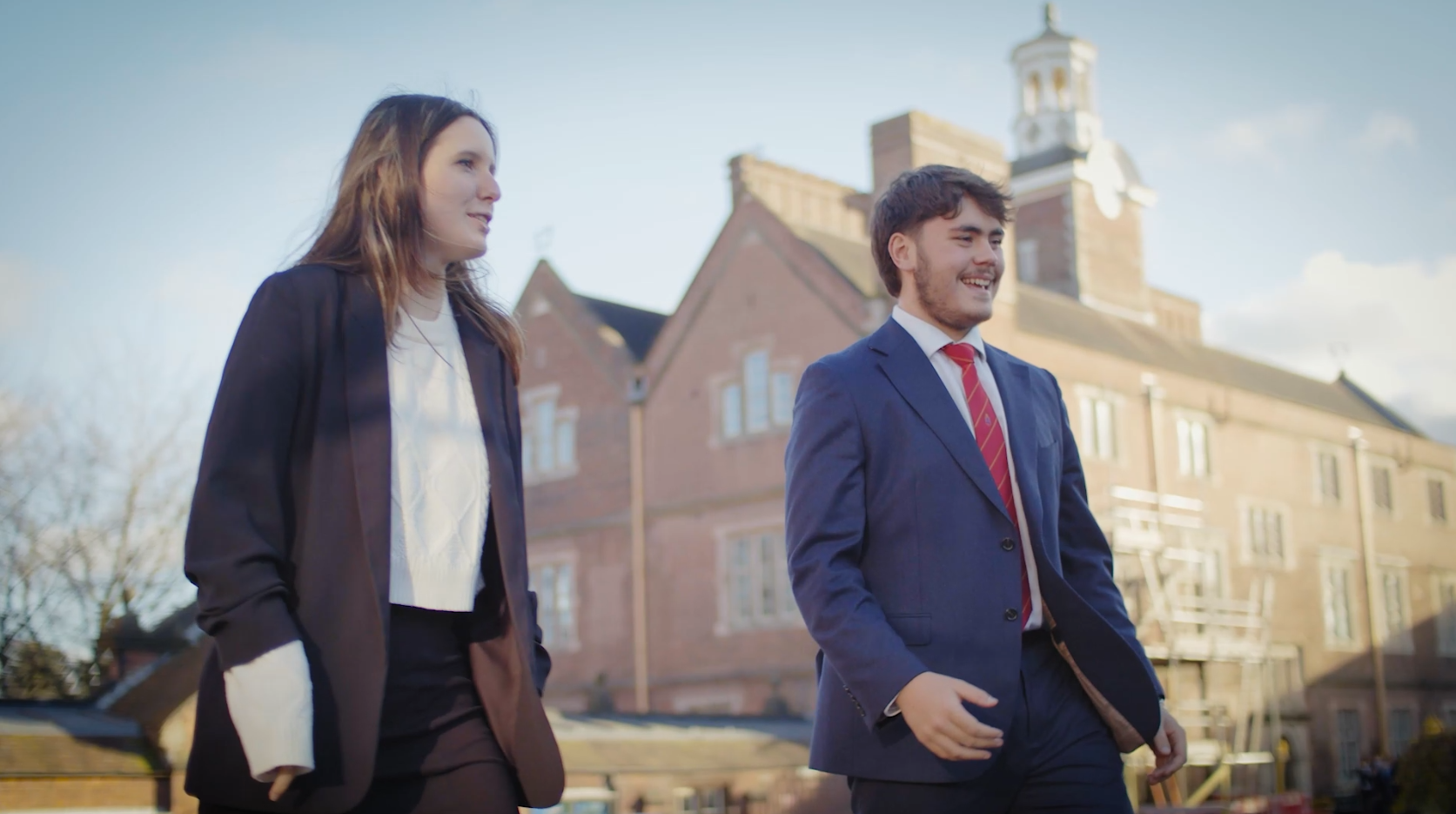olivia and noah walk side-by-side across school in beautiful, both dressed in suits with trees and founders building behind them