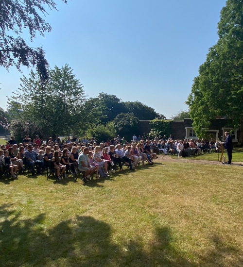 Prospective  students and families gather seated in a beautiful rose garden in summer sunshine, listening to the Headmaster's welcome address on open morning