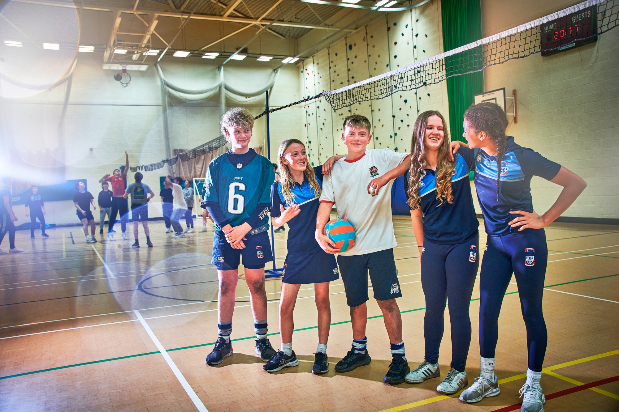 Five students, boys and girls, in the sports hall holding a volleyball, leaning on each other and chatting