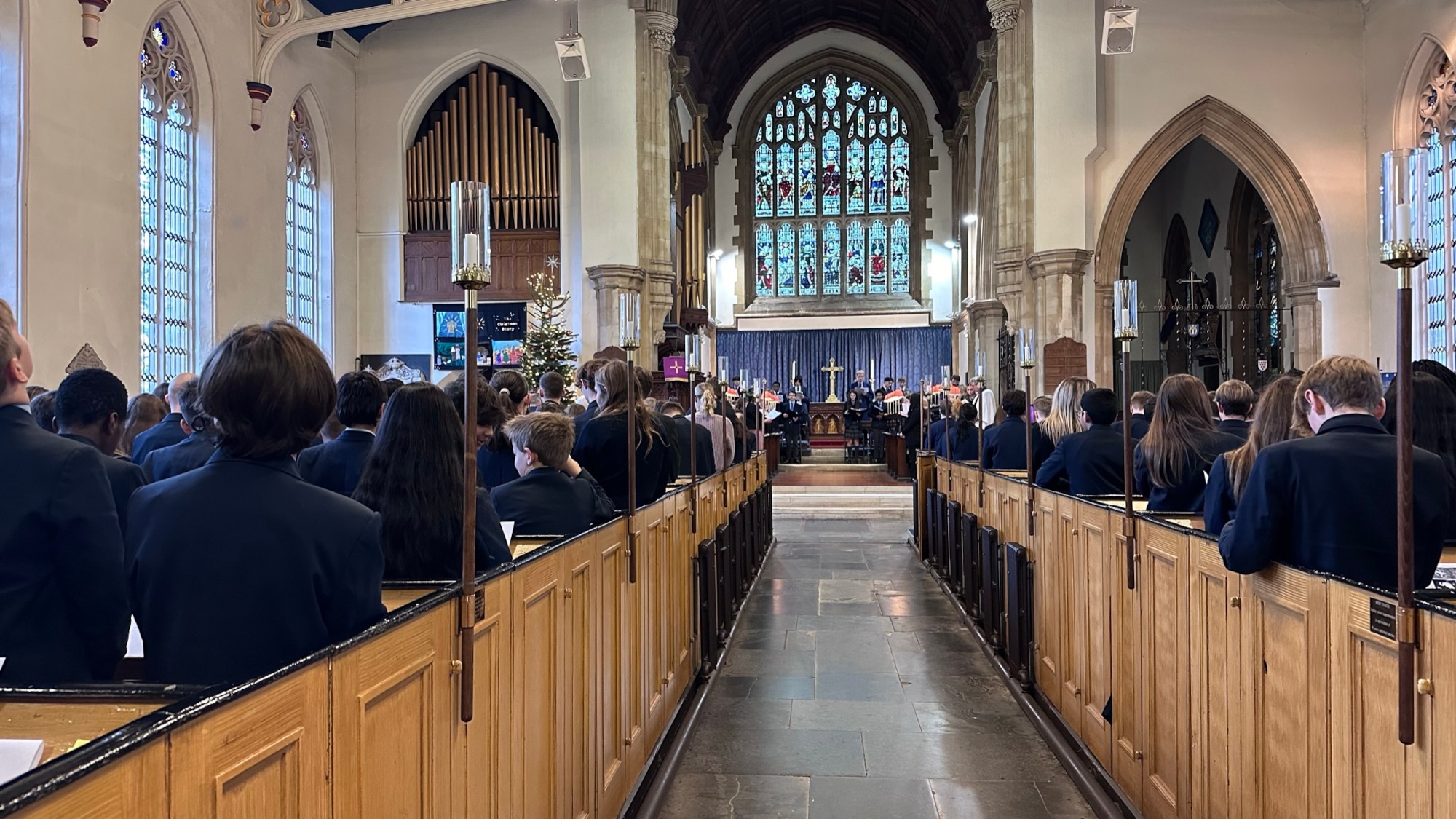 church pews filled with students of all ages with the camera looking straight down the isle to a beautiful stained glass window and choir standing facing the congregation