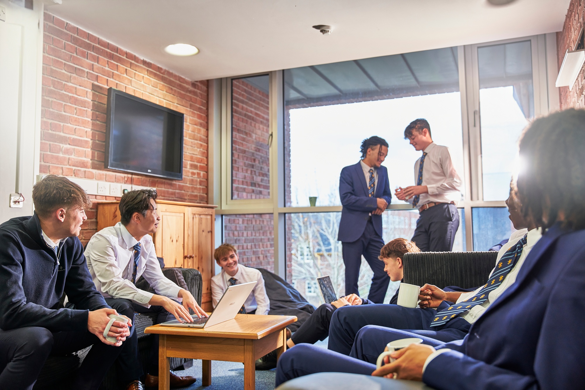 senior sixth form boy boarders sitting on sofas and the floor in common space in the boarding house, with a couple standing infront of a window overlooking campus