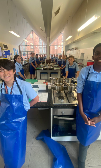 Year 7 students stand smiling in a teaching kitchen wearing aprons, ready to start cooking