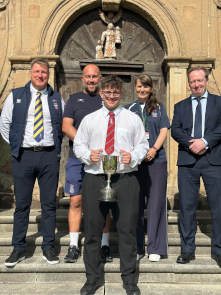 Student holding a silver cup stands with four members of staff smiling as he receives an award