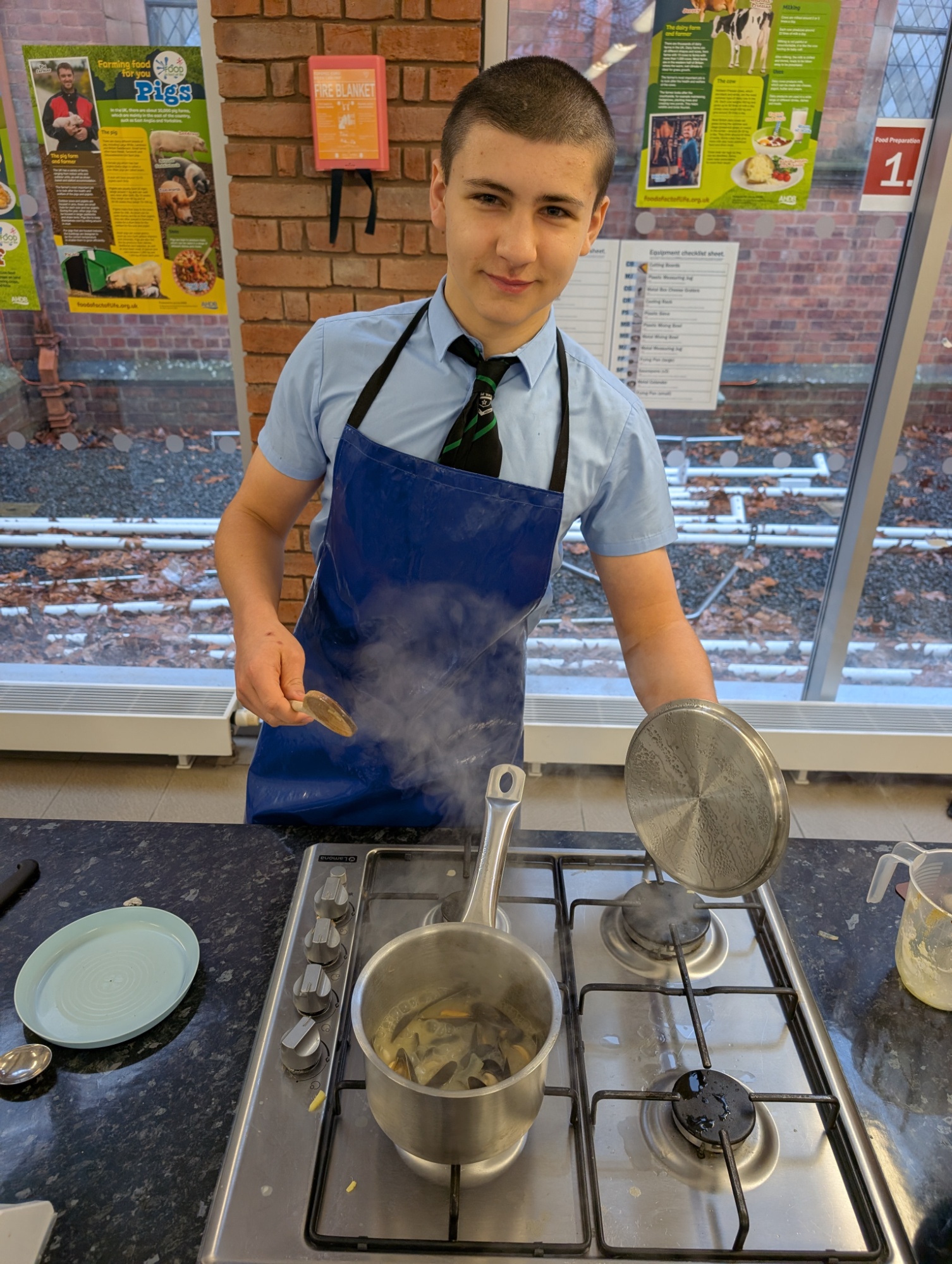 Student in a blue cooking apron watching mussels boiling on a stove