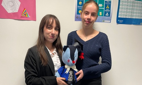 Two female sixth form students smiling and holding their team mascot, a blue teddy bear made of OSH uniform